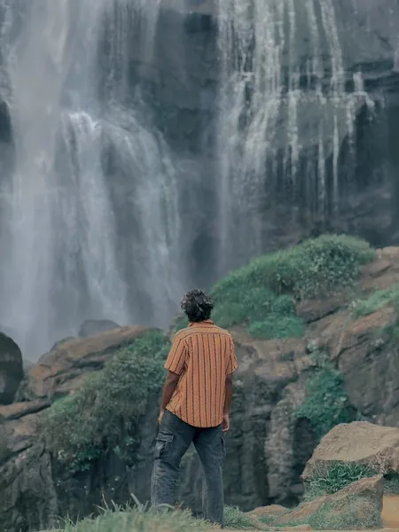 A man in a striped shirt, seen from behind, standing on rocks in front of a tall waterfall